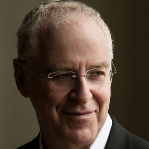 An older man with short, gray hair and glasses, wearing a white shirt and dark blazer, looks to the side with a slight smile against a dark, neutral background.