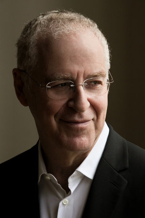 An older man with short, gray hair and glasses, wearing a white shirt and dark blazer, looks to the side with a slight smile against a dark, neutral background.