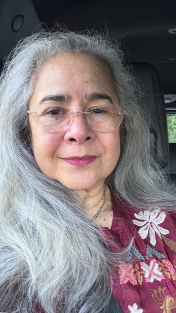 A woman with long gray hair and glasses is smiling slightly. She is wearing a maroon top with floral embroidery and is seated in a car.