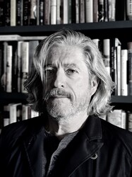 A middle-aged man with shoulder-length gray hair and a beard stands in front of a bookshelf filled with books. He wears a dark jacket and looks at the camera with a neutral expression.