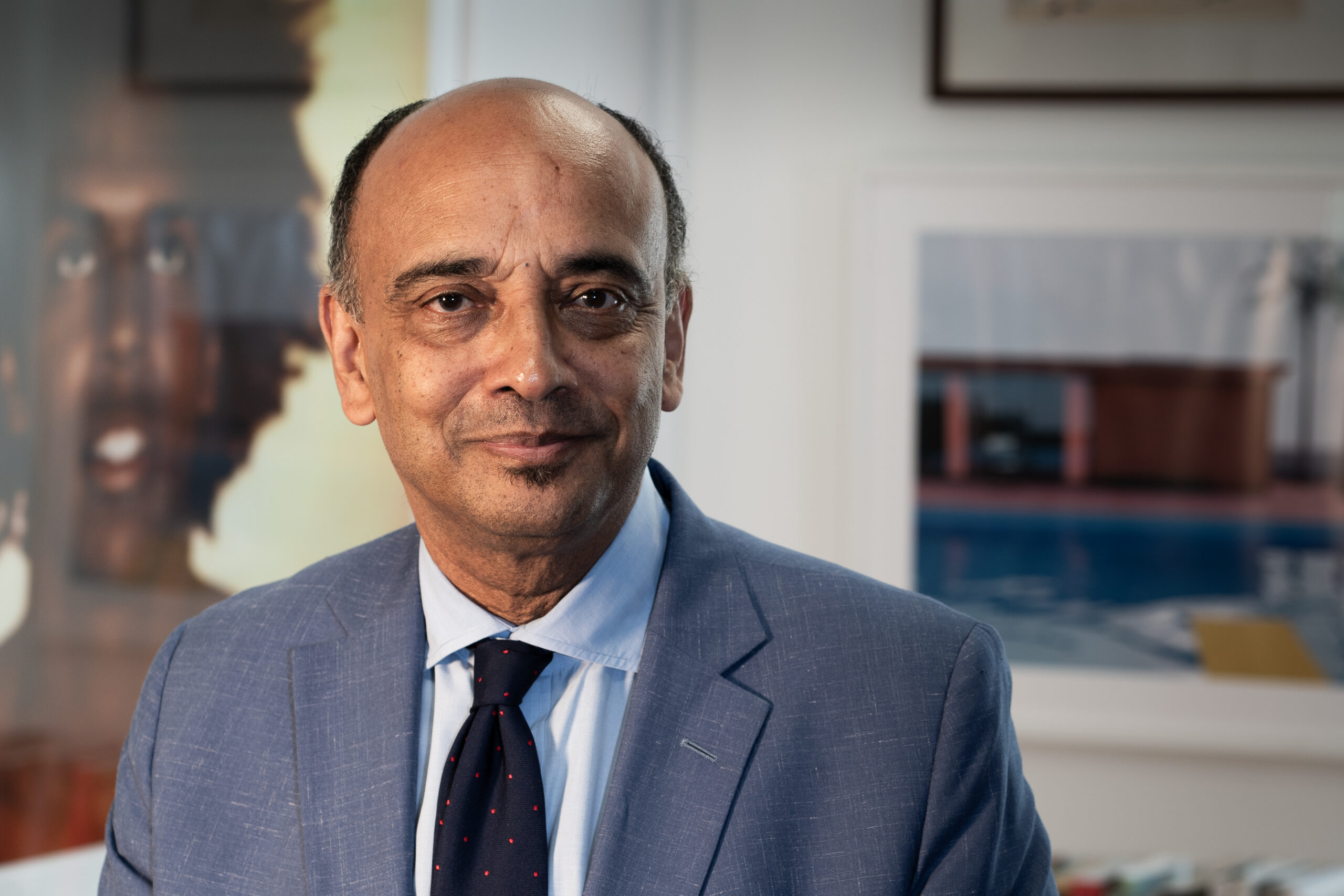 A middle-aged man in a blue suit and tie smiles slightly, standing in a well-lit office with artwork and framed photographs in the background.