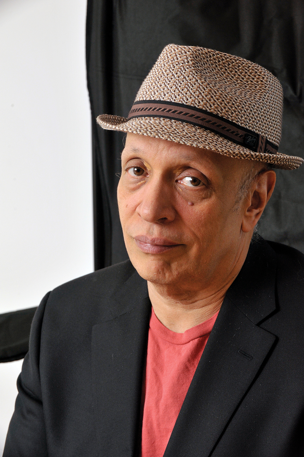 A man wearing a black blazer, red shirt, and a woven fedora hat looks at the camera with a neutral expression. The background is simple, featuring black and white areas.