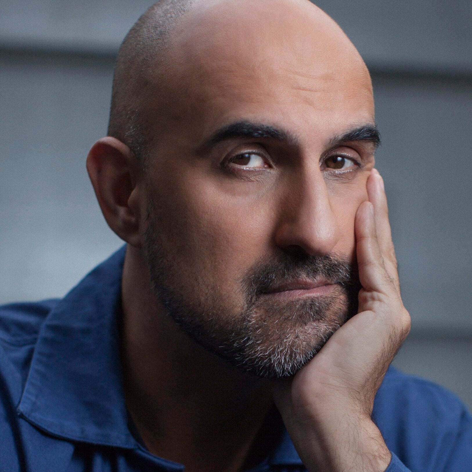 A bald man with a trimmed beard rests his face on his hand, looking thoughtfully at the camera. He wears a blue jacket over a striped shirt, with a neutral gray background.