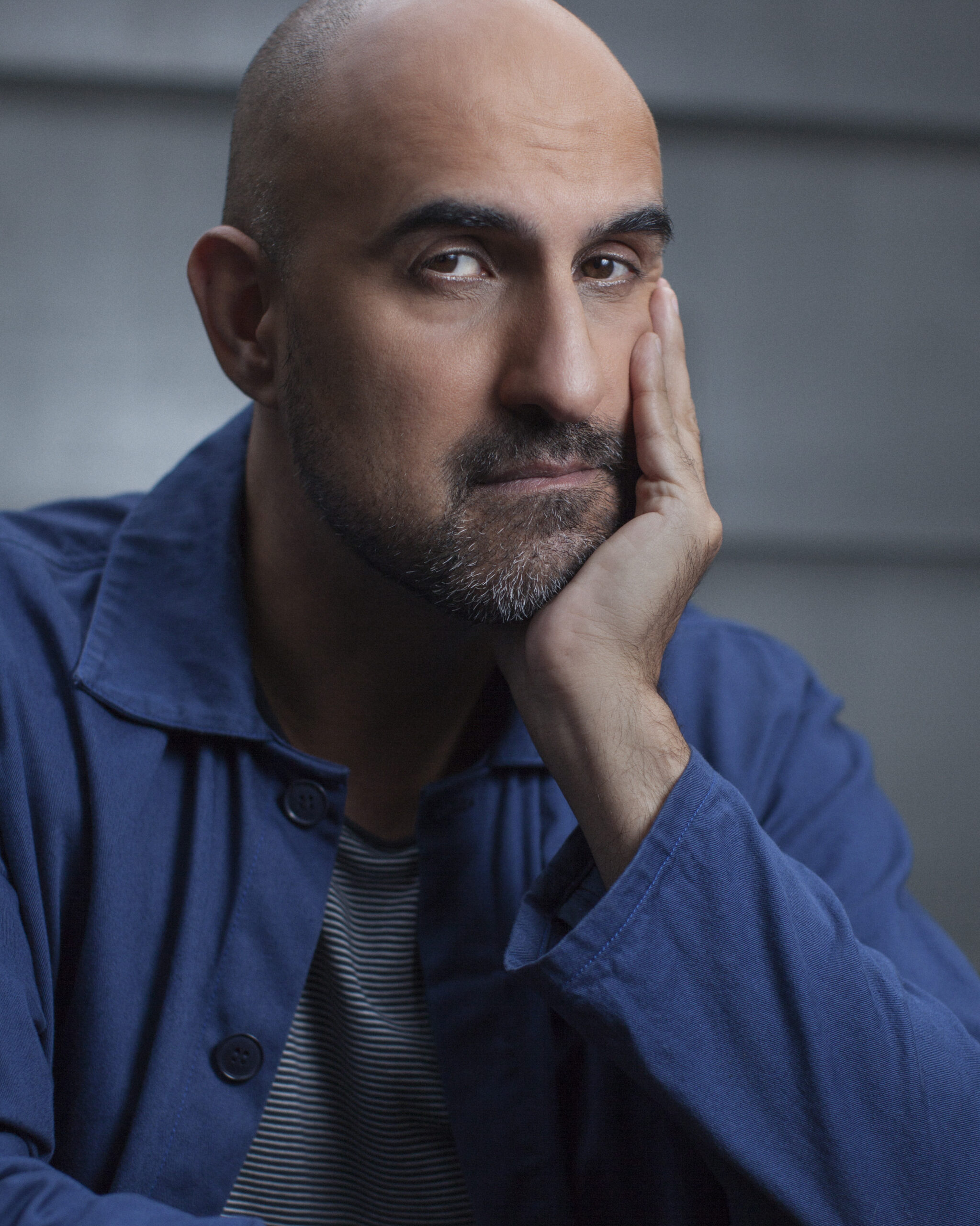 A bald man with a trimmed beard rests his face on his hand, looking thoughtfully at the camera. He wears a blue jacket over a striped shirt, with a neutral gray background.