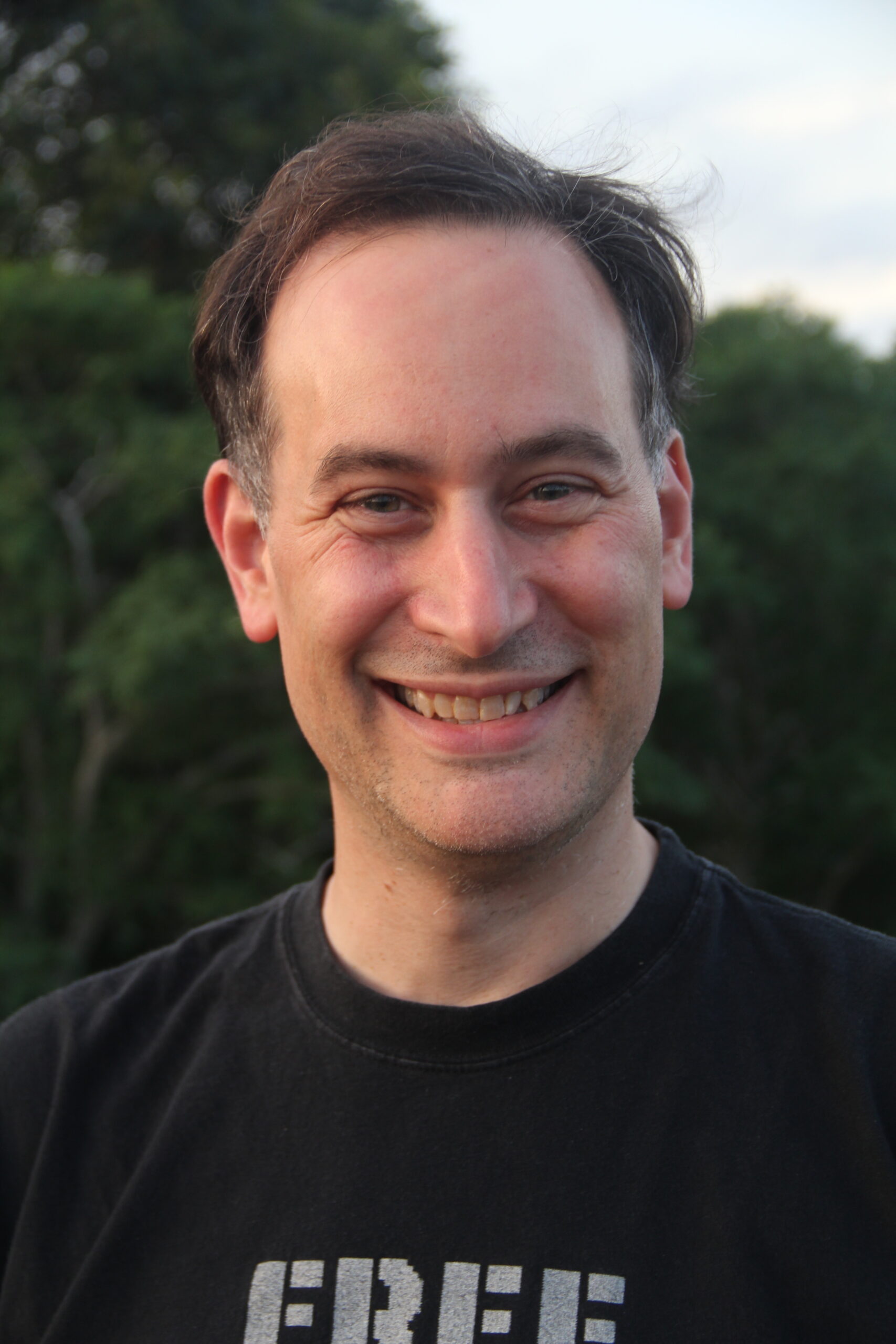 A man with short dark hair and a receding hairline smiles at the camera. He is wearing a black T-shirt and standing outdoors with green trees in the background.