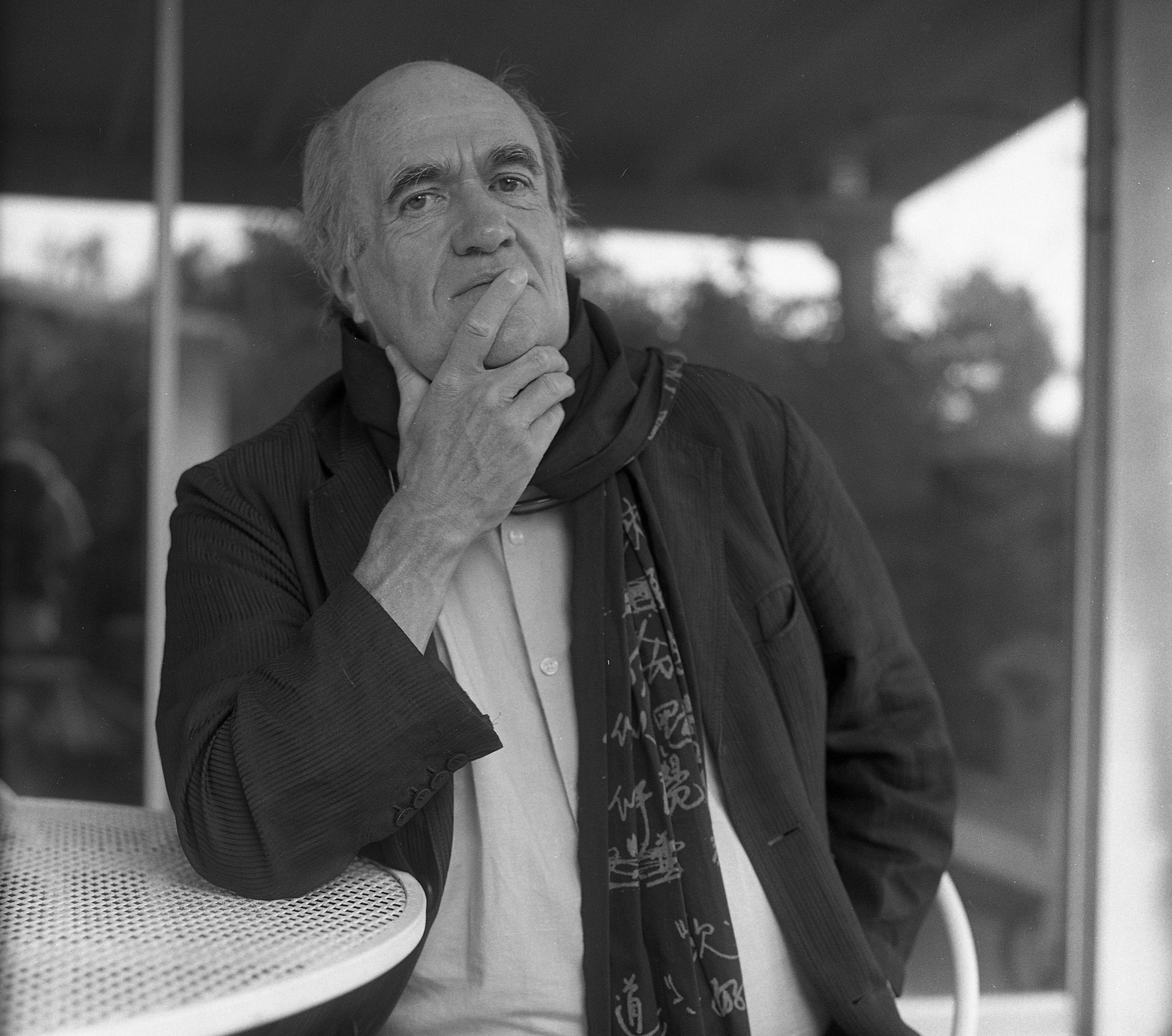 Elderly person with a thoughtful expression, wearing a dark jacket, light shirt, and a patterned scarf, sits at a round table with a hand on their chin. The background features a blurred outdoor area with trees and a building structure.