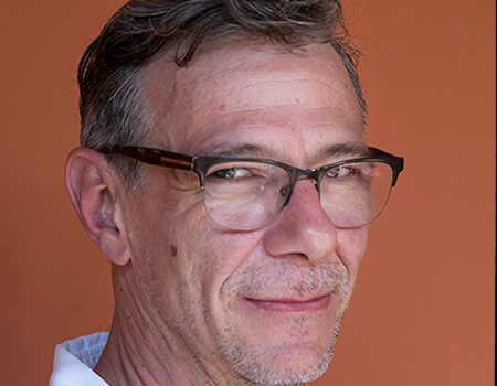 A middle-aged man with short brown hair, glasses, and slight stubble smiles subtly at the camera. He is wearing a white shirt and standing in front of an orange background.