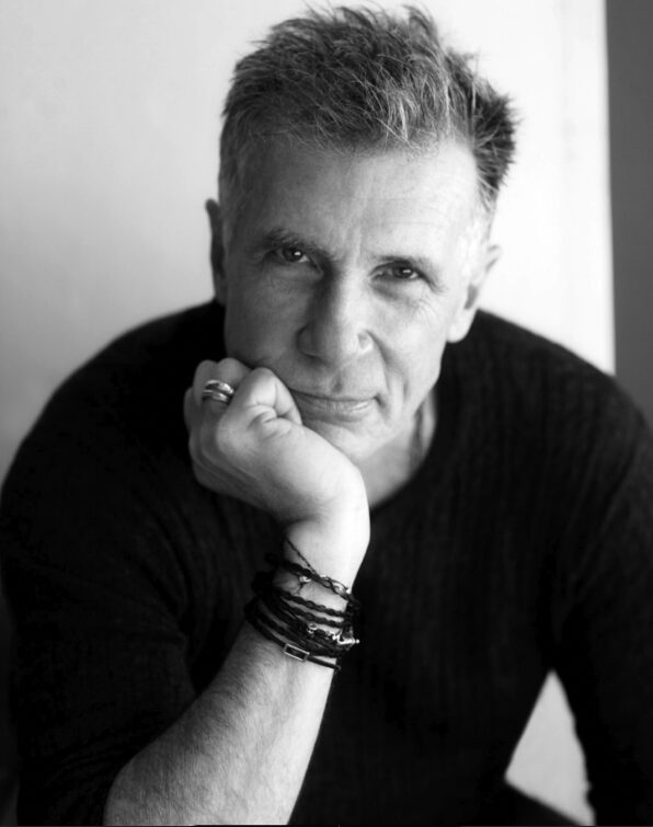 A grayscale photo of a middle-aged person with short, textured hair, resting their chin on their fist. They wear several bracelets and rings, and have a thoughtful expression. The background is simple and out of focus.