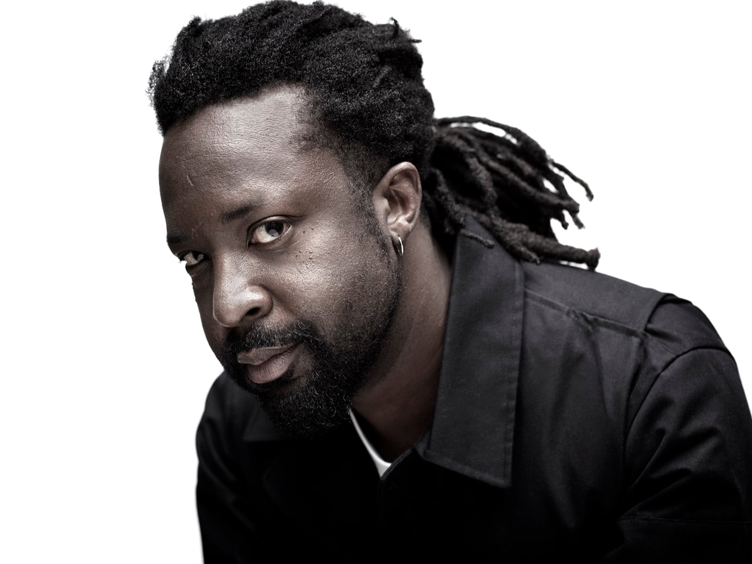 A man with medium-dark skin, black dreadlocks, and a short beard looks at the camera with a slight smile. He wears a black jacket and a small hoop earring, set against a plain white background.