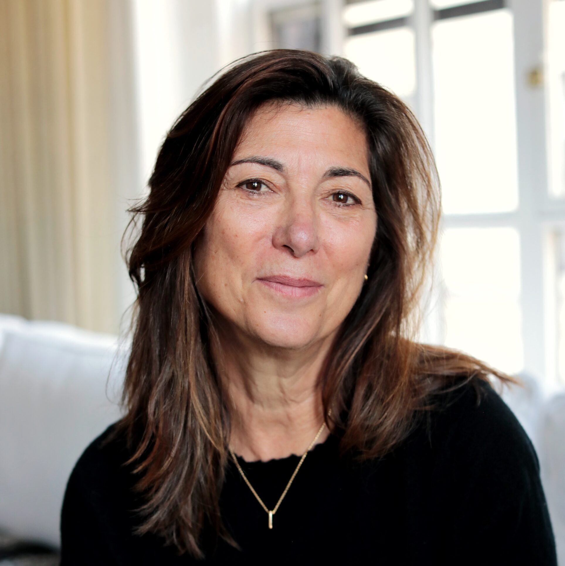 A woman with medium-length brown hair, wearing a black top and a gold necklace, sits indoors in front of a window with white curtains, softly smiling at the camera.