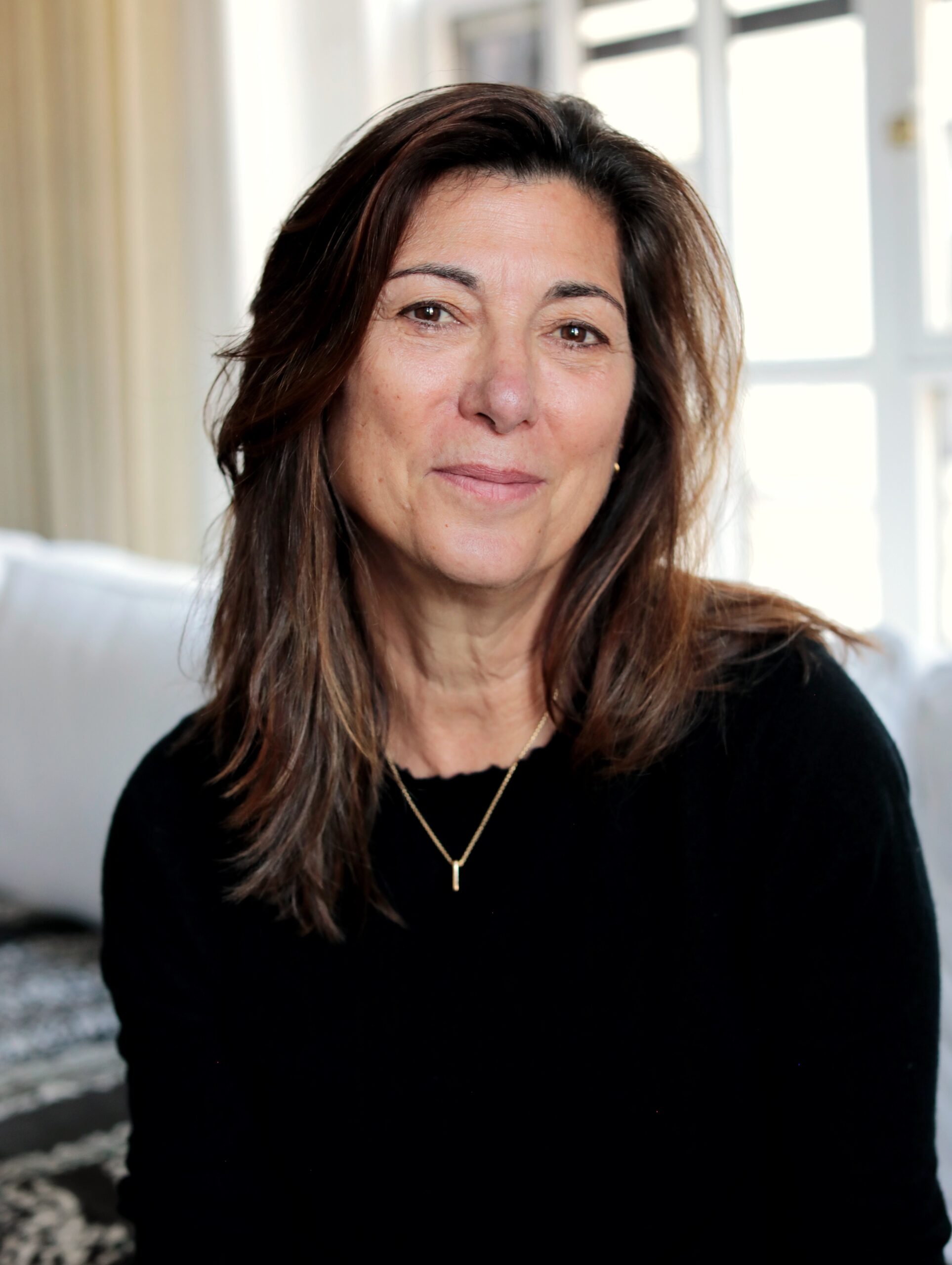 A woman with medium-length brown hair, wearing a black top and a gold necklace, sits indoors in front of a window with white curtains, softly smiling at the camera.
