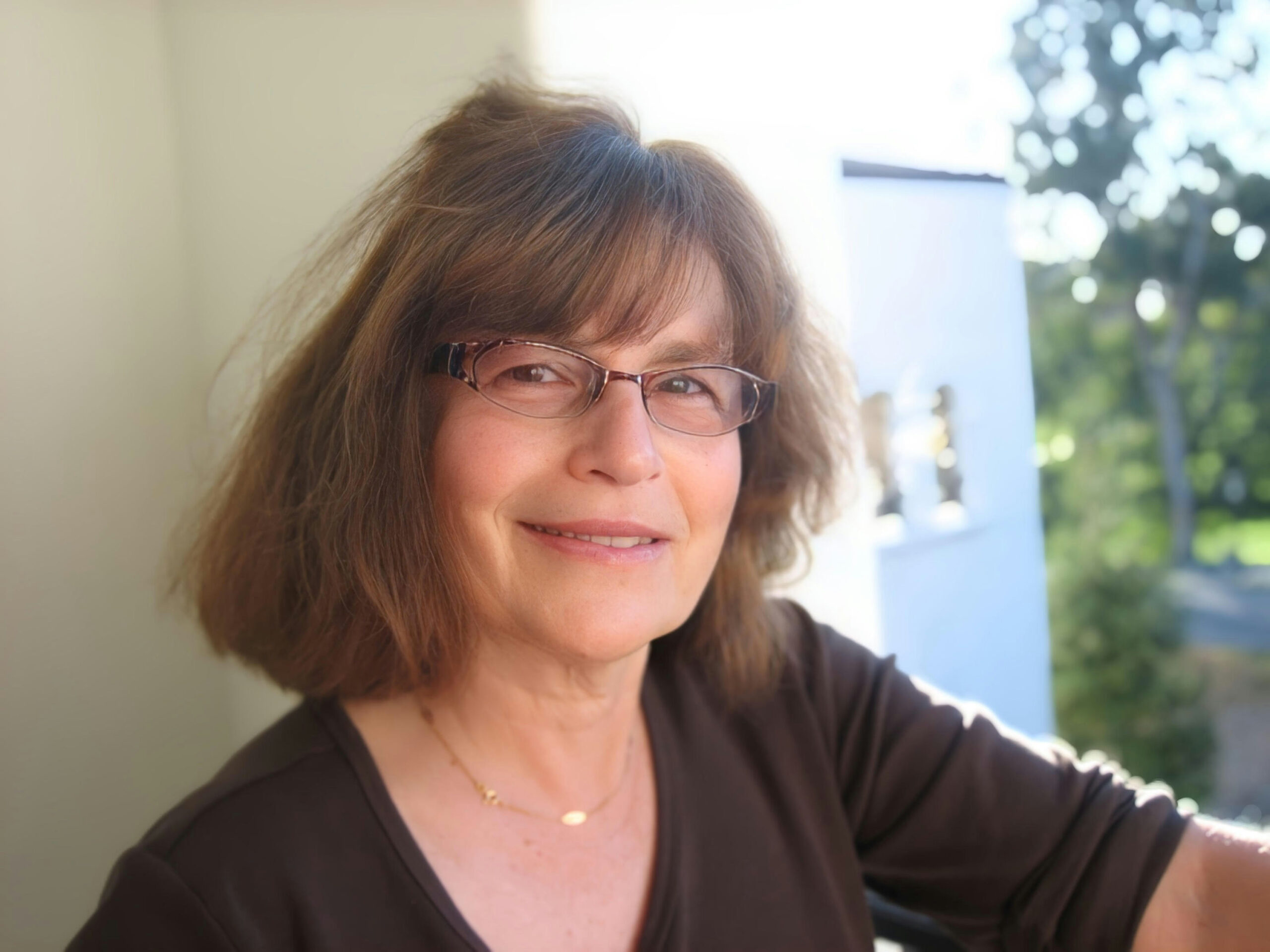 A woman with shoulder-length brown hair and glasses smiles at the camera. She is wearing a brown top and a delicate necklace. The background is bright and sunny, with some greenery visible outside.
