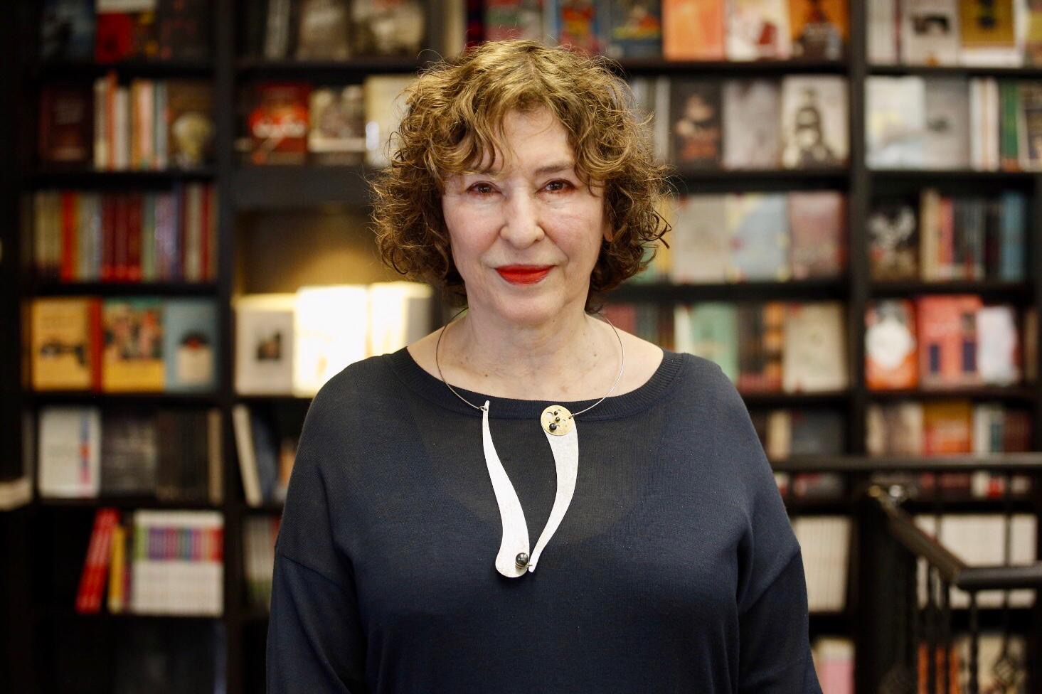A woman with curly hair and red lipstick stands in front of bookshelves filled with books. She is wearing a dark top and a distinctive white necklace. The background is softly blurred, focusing attention on her.