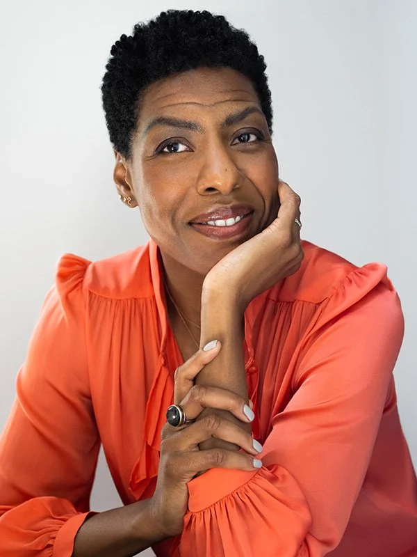 A woman with short curly hair wearing a coral blouse smiles gently, resting her chin on her hand. She has light nail polish, a black ring, and sits against a plain, light background.