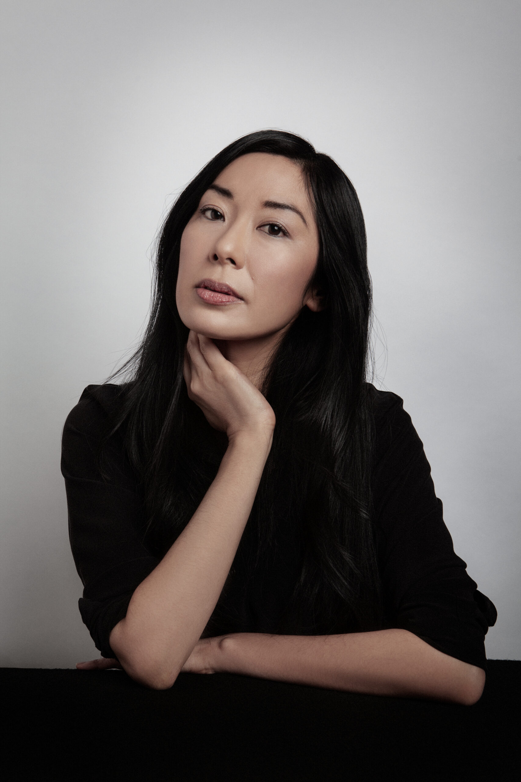 A woman with long black hair sits at a table against a plain background, resting her chin on her left hand. She wears a black top and looks thoughtfully at the camera.