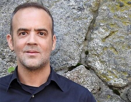 A man with short hair and a trimmed beard, wearing a dark collared shirt, stands in front of a textured stone wall with patches of moss.