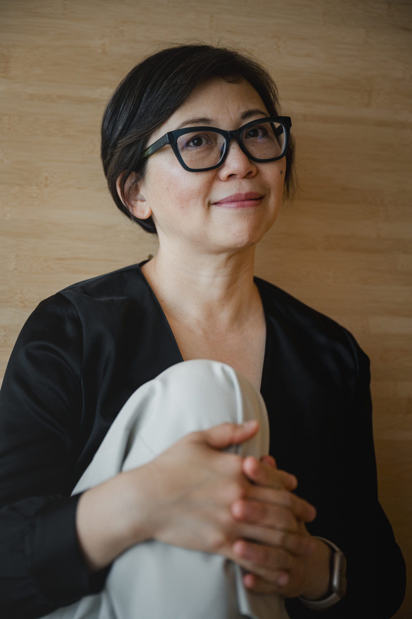 A woman with short dark hair and glasses, wearing a black blouse and light pants, sits indoors with her hands clasped around her knees, looking up and smiling softly against a wooden background.