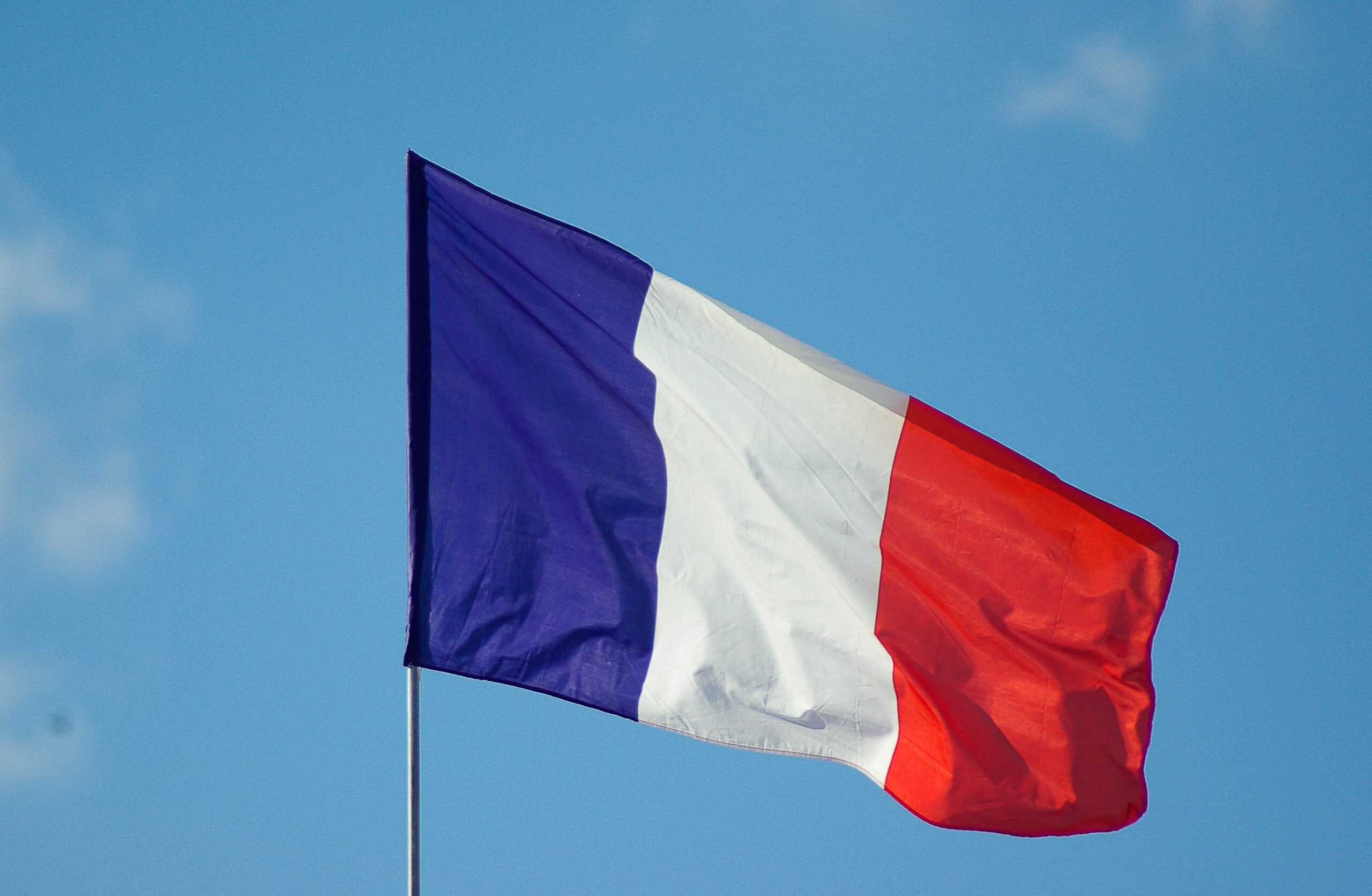 The image shows the French flag waving against a clear blue sky. The flag has three vertical stripes: blue on the left, white in the middle, and red on the right.