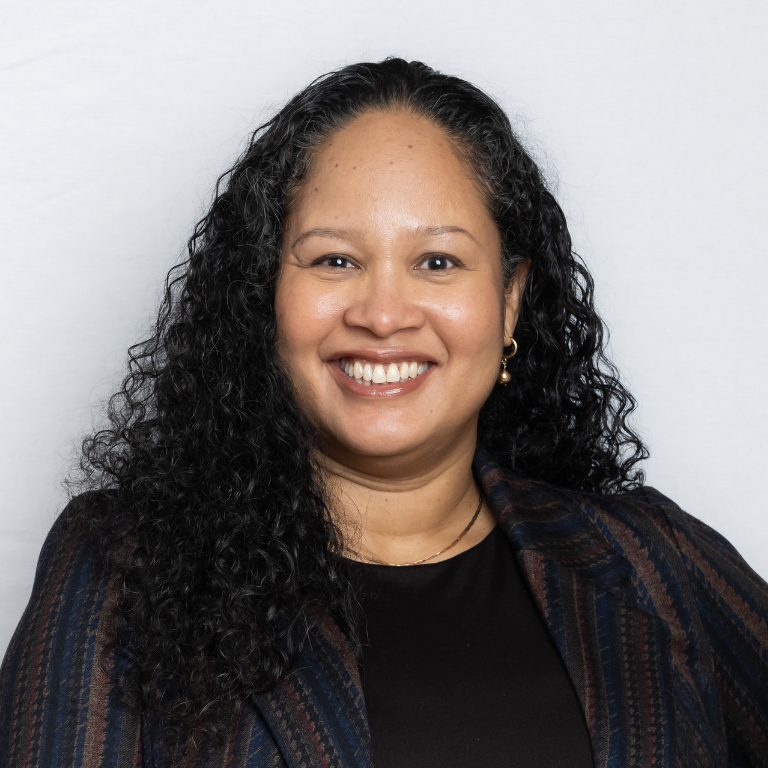 A woman with long curly hair, wearing a striped blazer over a black top, smiles at the camera against a plain light background.