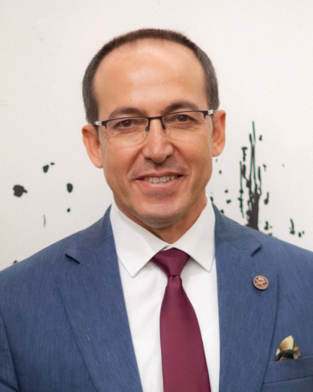 A man wearing glasses, a blue suit, white shirt, and maroon tie, smiling in front of a white background with black paint splatters.