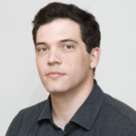 A person with short dark hair is wearing a dark gray collared shirt and looking at the camera against a plain light background.