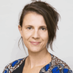 A person with short, styled hair and a neutral expression is posing against a plain background. They are wearing a patterned top with blue and brown details.