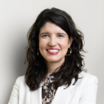 A woman with long, wavy dark hair, wearing a white blazer and patterned blouse, smiles at the camera against a plain light-colored background. She is also wearing gold hoop earrings.