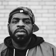 A man with a beard wearing a hoodie, jacket, and a baseball cap turned backward stands in front of a brick wall, looking directly at the camera. The photo is in black and white.