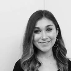 A young woman with long wavy hair, wearing hoop earrings and a dark top, smiles at the camera in front of a plain light-colored background. The image is in black and white.