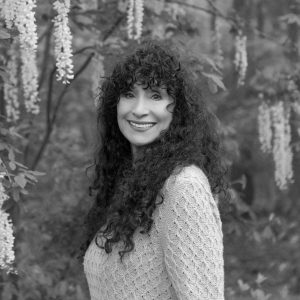 A woman with long, curly dark hair smiles at the camera while standing outdoors among hanging flowers and leafy branches. She is wearing a textured sweater. The black-and-white photo has the elegance of a Pen America Literary Awards portrait.