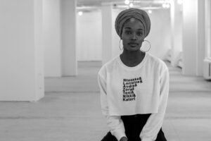 A young woman wearing a headwrap and hoop earrings sits in a spacious, light-filled room. She wears a theater award sweatshirt and looks calmly at the camera. The image is in black and white.