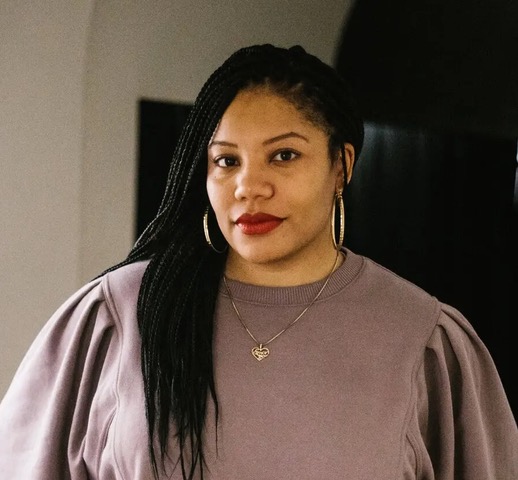 A woman with long braided hair, wearing hoop earrings, a necklace with a heart pendant, and a mauve sweater, stands indoors against a dark background, looking at the camera with a slight smile—her poise hinting at a recent theater award.