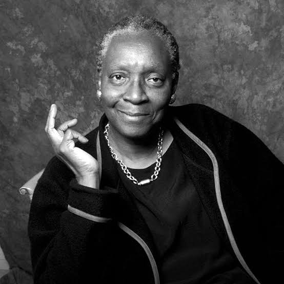A smiling person with short hair and a dark outfit sits against a textured background, evoking an air of artistic elegance reminiscent of a Nabokov Award portrait. One hand is raised in a relaxed manner, and they wear a necklace. The image captures the timeless charm in black and white.