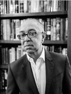 Black and white photo of an older man wearing glasses and a suit, standing in front of a bookshelf filled with books. He has a neutral expression and looks directly at the camera, evoking the dignified presence of a Mike Nichols award recipient.