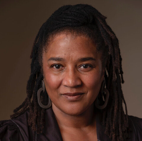 A woman with medium-dark skin and shoulder-length locs, wearing large hoop earrings and a dark, collared top, looks confidently at the camera against a plain brown background.