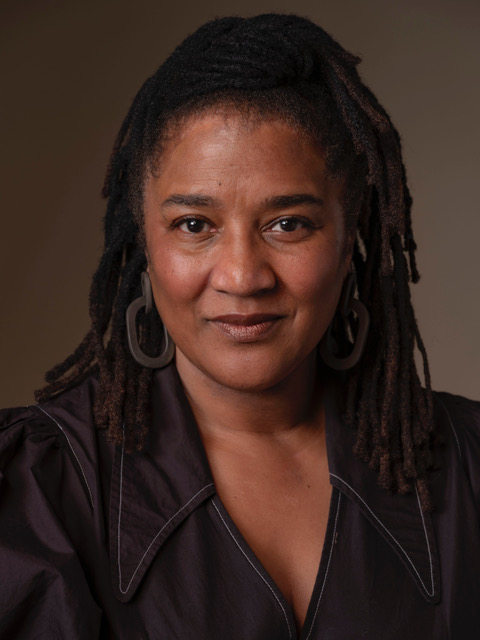 A woman with medium-dark skin and shoulder-length locs, wearing large hoop earrings and a dark, collared top, looks confidently at the camera against a plain brown background.