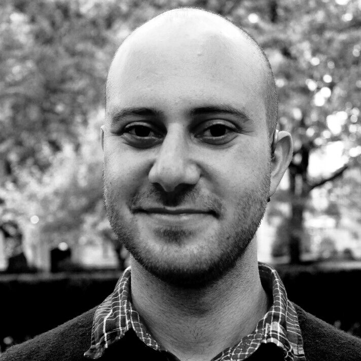 A bald man with a short beard and slight smile is wearing a plaid shirt. He stands outdoors before blurred trees in this black and white image, reminiscent of portraits of PEN America Literary Awards judges 2021.