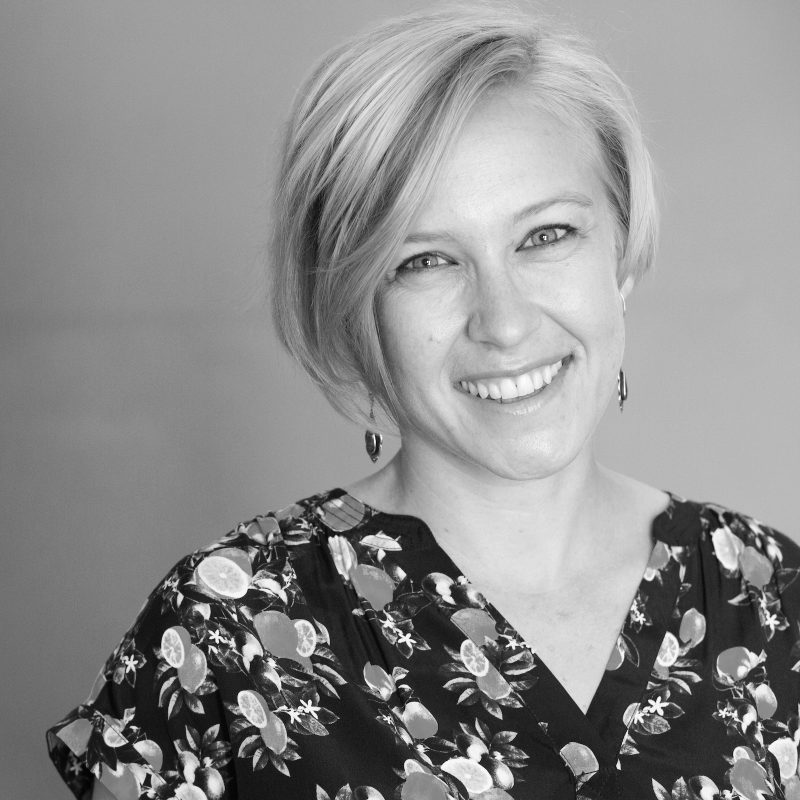 A woman with short blonde hair, one of the PEN America Literary Awards judges 2021, smiles at the camera. She is wearing a patterned blouse with fruit designs and earrings. The black and white photo features a plain background.