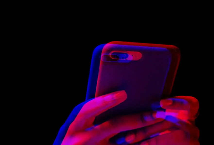 A pair of hands holds a smartphone against a black background, pulsing with a vibrant red and blue 3D effect, echoing the chaotic digital landscape where social media can sometimes foster online abuse.