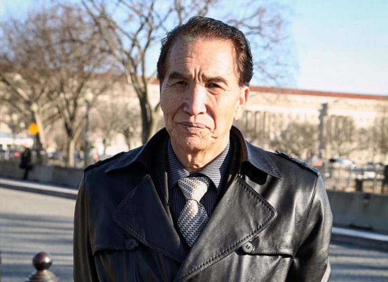 A man with dark hair wearing a black leather jacket, shirt, and patterned tie stands outdoors on a sunny day, with leafless trees and a large building in the background—a scene reminiscent of the spirit behind the Freedom to Write Index 2020.