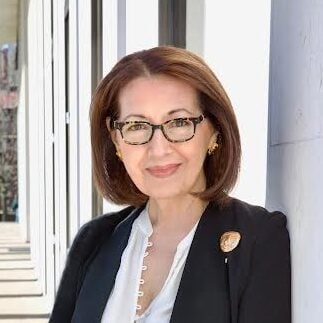 A woman with shoulder-length brown hair, wearing glasses, a black blazer, and a white blouse, stands smiling against a white wall in a sunlit corridor with columns.