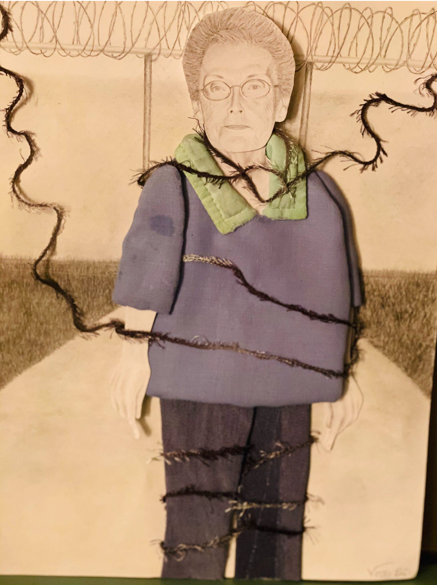 Textile display of a woman standing in front of a barbed wire fence drawn in pencil. She is wearing a purple sweater composed of fabric with a black thread wrapped around her limbs.