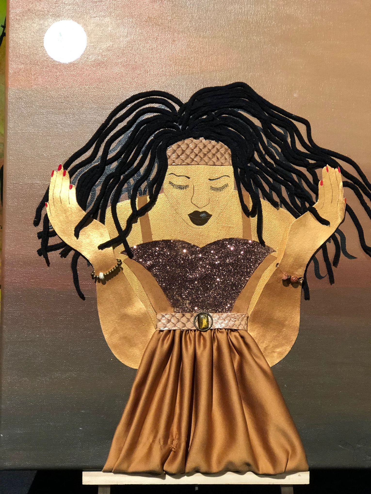 A woman with long locs kneels in prayer with her eyes closed and hands faced upwards. She is wearing a glitter top and a gold satin skirt.