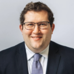 A person with short curly hair and glasses is smiling at the camera. They are wearing a dark suit, a white shirt, and a patterned tie. The background is a plain light gray.