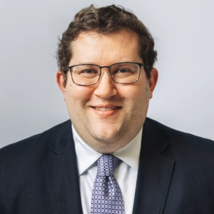 A person with short curly hair and glasses is smiling at the camera. They are wearing a dark suit, a white shirt, and a patterned tie. The background is a plain light gray.