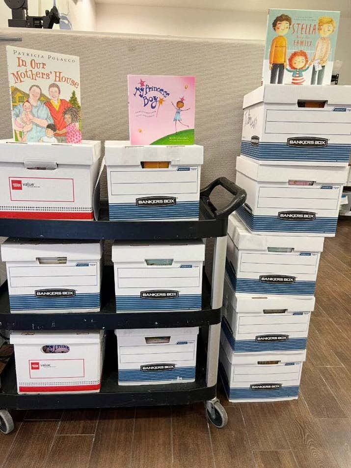 A cart and stack of white storage boxes on a wooden floor, with three banned books for children displayed upright on top, including In Our Mothers’ House, The Princess Dog, and Stella Brings the Family.