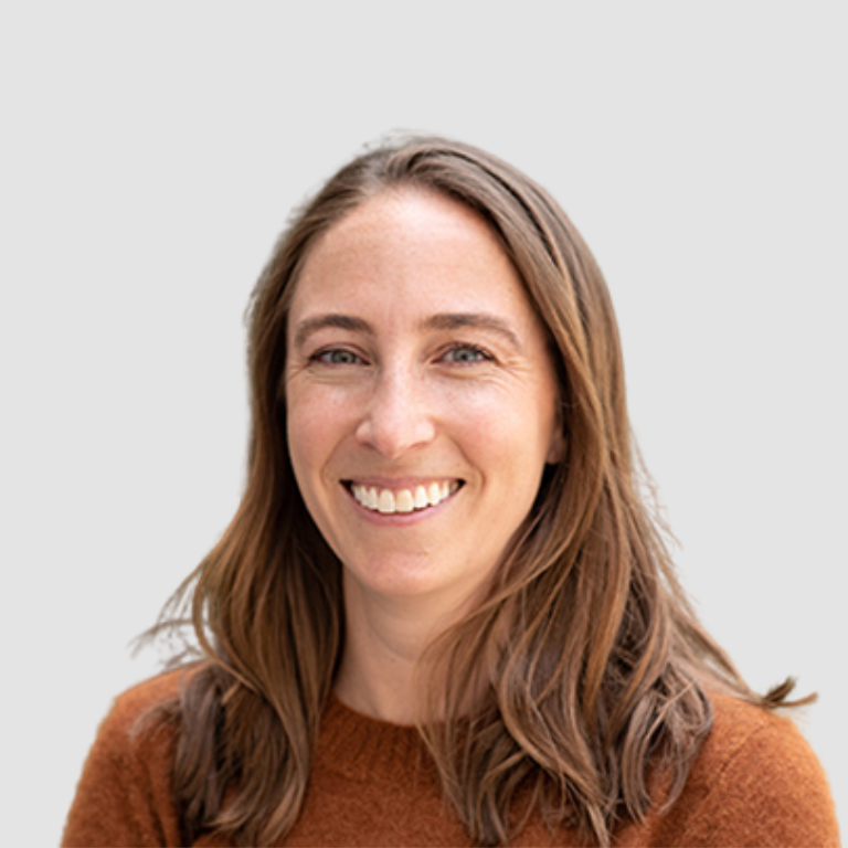 A woman with long brown hair, wearing a rust-colored sweater, smiles at the camera against a plain light gray background.