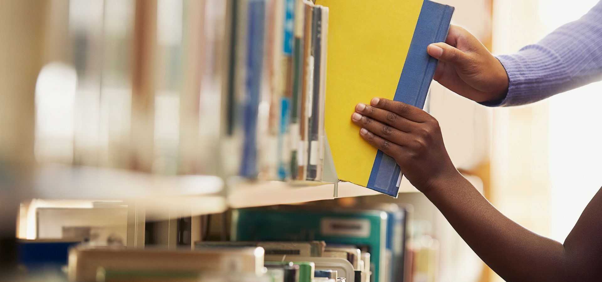 A close-up of two hands reaching for books on a library shelf—one holding a yellow book with a blue spine—captures the essence of Pen America’s commitment to reading between the lines. Rows of books are visible in the background.