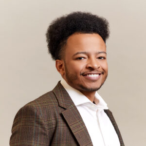 A man with short curly hair and a trimmed beard, wearing a brown plaid suit jacket over a white dress shirt, smiling in front of a plain light background.