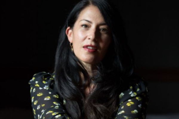 In the dim light, a person with long dark hair stands against a shadowy backdrop, adorned in a black top with floral patterns. The light falls just right, as if offering writing advice on how to highlight their features.